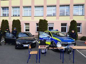 Po lewej stronie stoi nieoznakowane bmw. Obok przy otwartych drzwiach stoi funkcjonariusz ruchu drogowego. Po prawej stronie stoi oznakowany radiowóz marki kia. Pojazd malowanych w nowych barwach tj. srebrnej, zielonej, niebieskiej oraz białej. Przy tym radiowozie również stoi funkcjonariusz