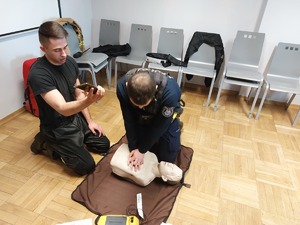 Policjant wykonuje masaż serca na fantomie, obok strażak dokonuje pomiaru korzystając z telefonu