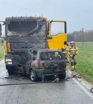 Na jezdni stoją pojazdy uczestniczące w zdarzeniu. Osobówka, która uderzyła w ciężarówkę uległa całkowitemu spaleniu. Samochód ciężarowy ma spaloną kabinę.