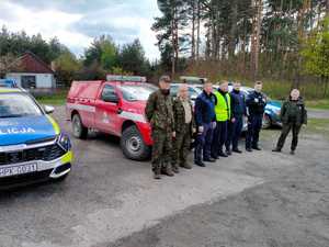 Na parkingu stoją funkcjonariusze policji oraz straży leśnej. Za nimi znajdują się radiowozy oraz pojazdy cywilne. W oddali widoczny las.