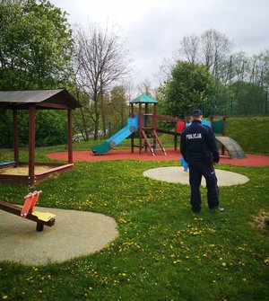 Dzielnicowy stoi na palcu zabaw. Zdjęcie przedstawia jego sylwetkę od tyłu. Na placu zabaw znajduje się konstrukcie w kształcie domków do zabaw.