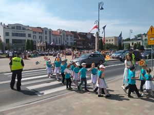 Dzieci przechodzą przez przejście dla pieszych. Na jednym z pasów jezdni stoją samochody.