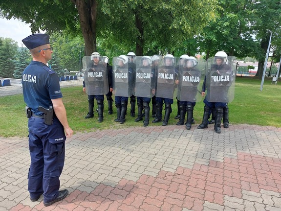 Policjanci umundurowani w  mundury ćwiczebne trzymając w ręku tarcze stoją dwu-szeregu są zwróceni w kierunku dowódcy drużyny.