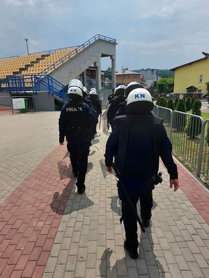 Policjanci ubrani w stroje ćwiczebne w kaski ochronne wyposażeni w tarcze i pałki idą chodnikiem znajdującym się na Stadionie Miejskim w Ropczycach.
