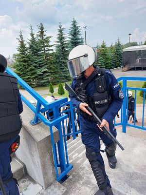 Na pierwszym planie funkcjonariusz który wchodząc po schodach na Stadionie Miejskim w Ropczycach trzyma w ręku strzelbę gładkolufową w oddali widoczni Kolejni funkcjonariusze którzy również wchodzą po schodach.