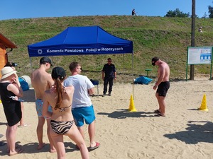 Jedna dorosła osoba pokonuje tor przeszkód miedzy pachołkami mając założone alkogoogle. Obok stoją inne osoby. W tle widoczny namiot z napisem policja