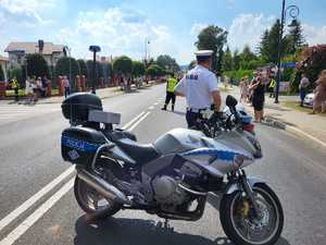 Na pierwszym panie stoi policyjny motocykl. Za nim widać funkcjonariusza a w oddali grupę ludzi idącą ulicą